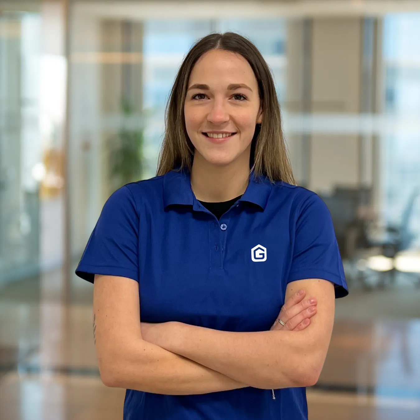 Smiling woman with crossed arms wearing a blue polo shirt with a white G logo in an office setting.