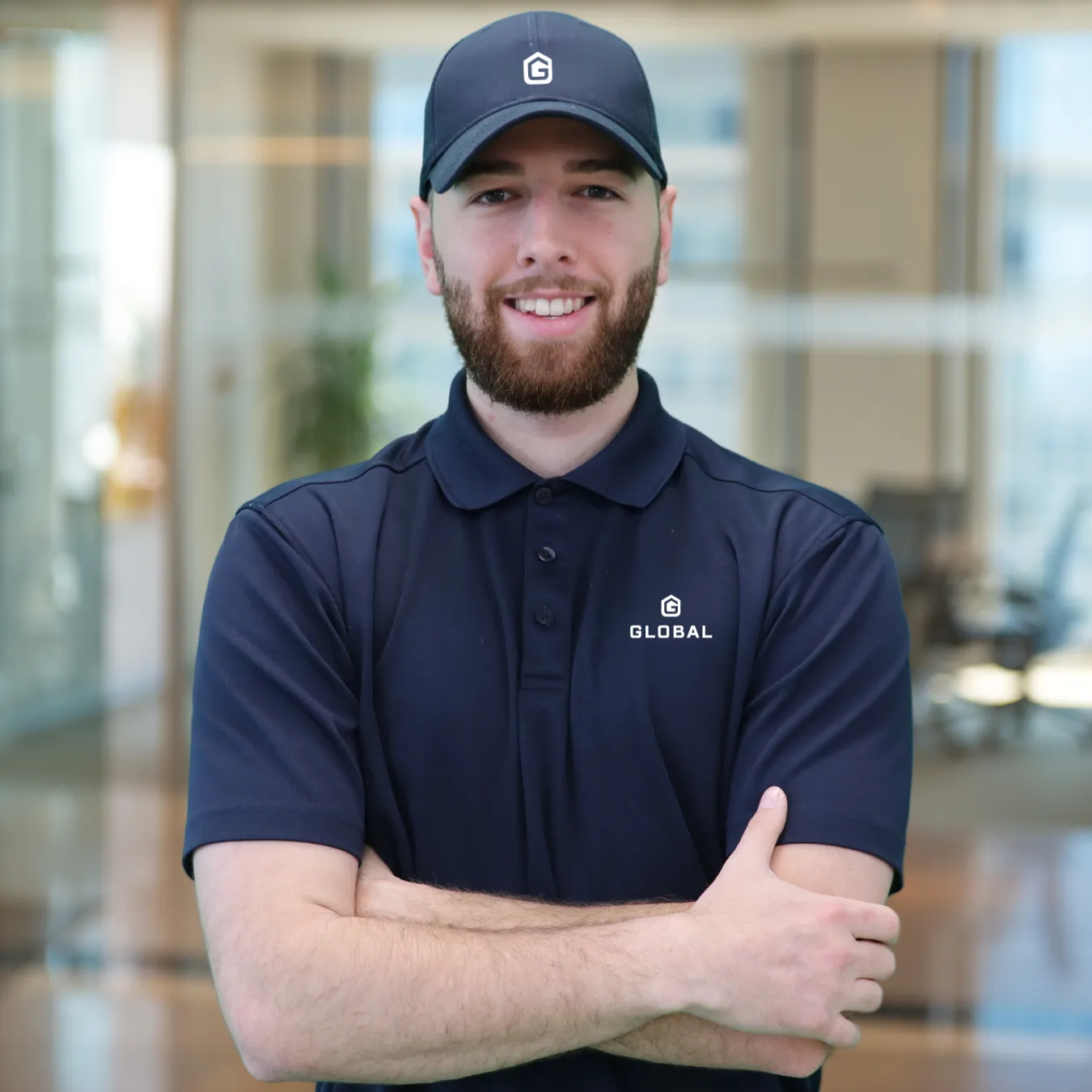 Man with beard wearing a navy polo shirt and cap with Global logo, standing with arms crossed in a modern office.