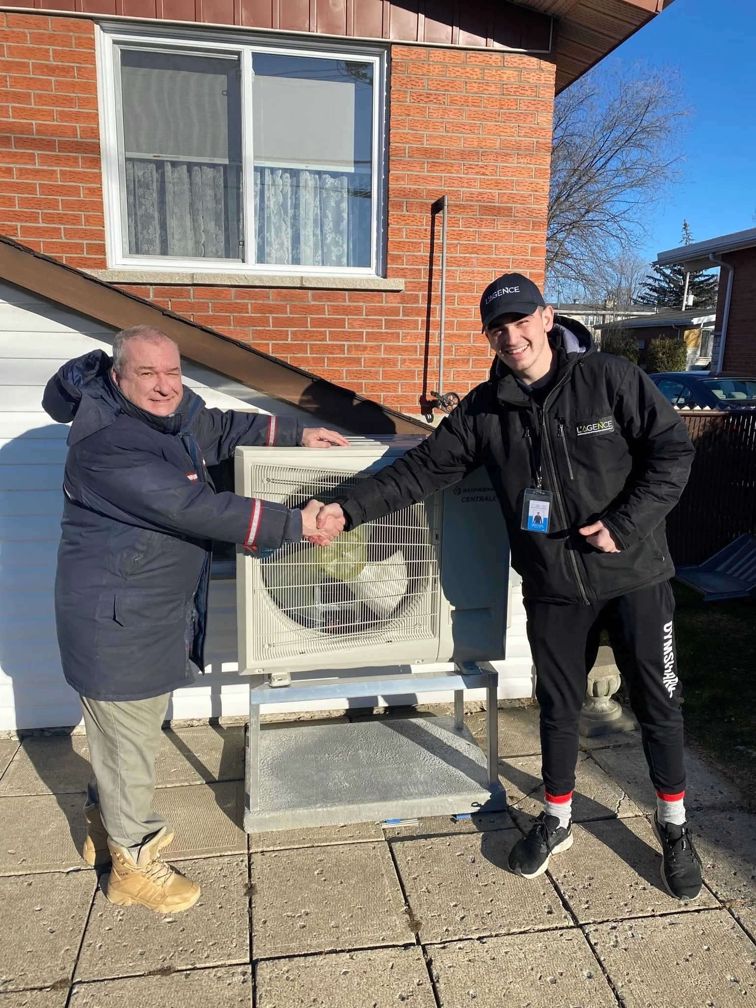Two men shaking hands outdoors in front of an HVAC unit mounted on a metal stand against a brick and white siding house.