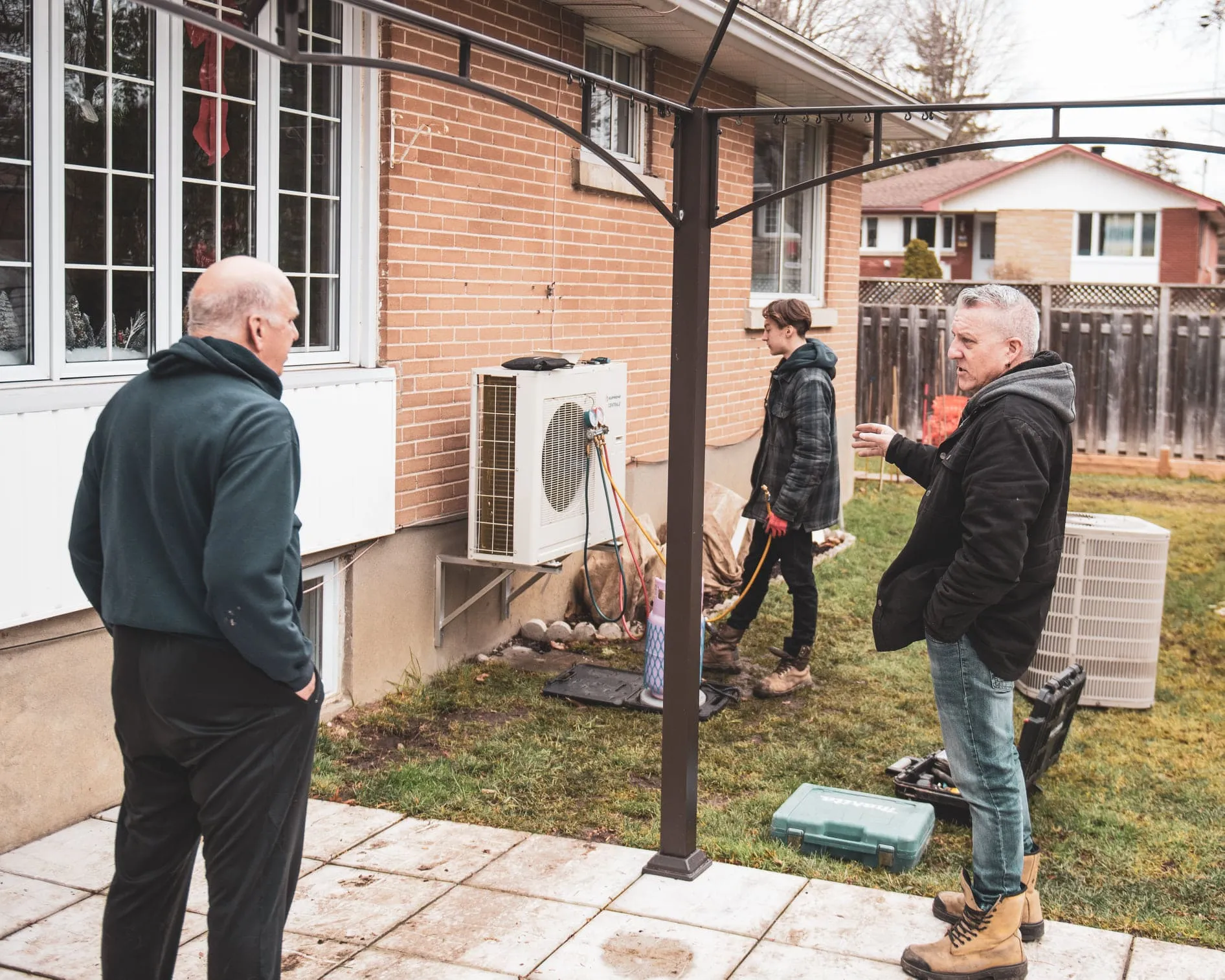 Three men working on or inspecting an air conditioning unit outside a brick house in a backyard.